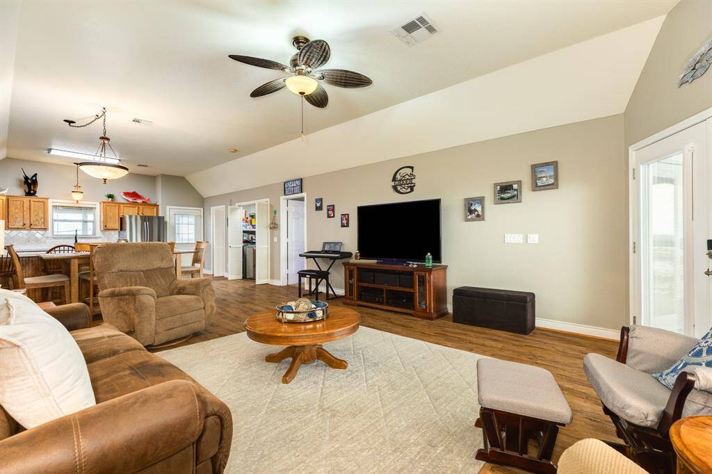 This spacious open-concept living area features a cozy sitting space with plush seating, a large flat-screen TV, and a ceiling fan. The room seamlessly flows into a modern kitchen with wooden cabinets, stainless steel appliances, and a dining space, all highlighted by warm wood flooring. Perfect for family gatherings and entertaining.