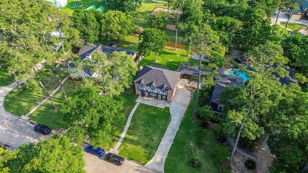 This aerial view showcases a charming Tudor-style home with a spacious driveway and lush green yard, surrounded by mature trees. Located in a tranquil neighborhood, it features a private backyard with gate access to the community park & pool!