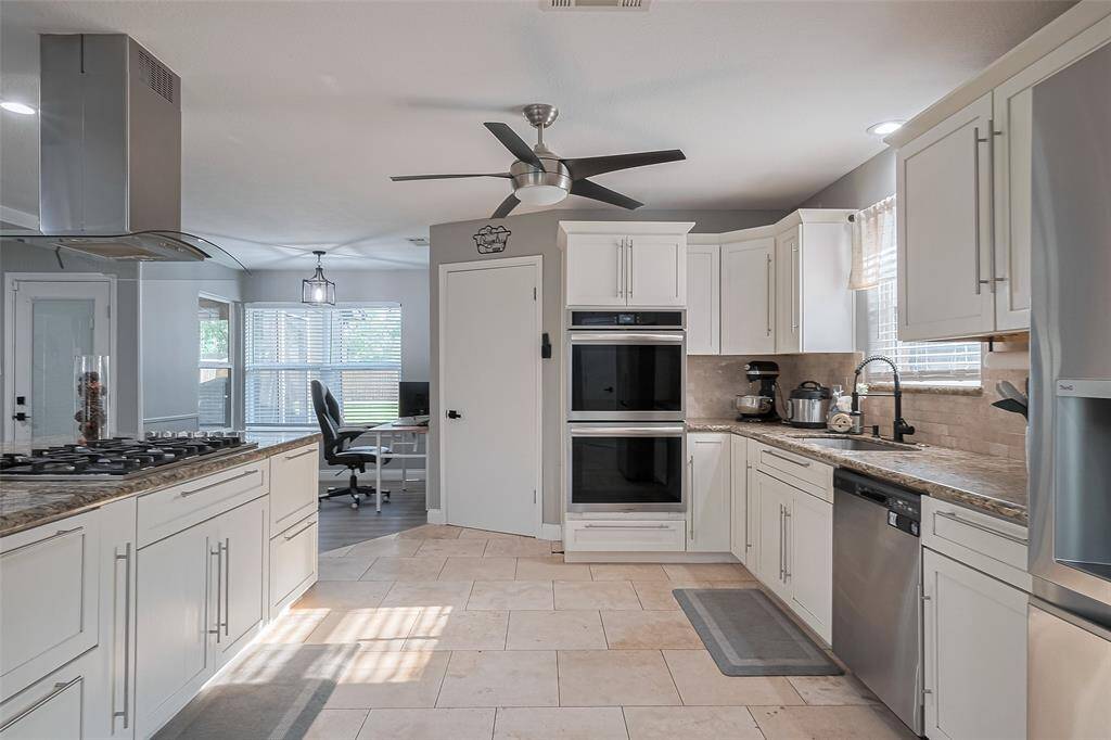 This modern kitchen features sleek white cabinetry, stainless steel appliances, and a spacious layout with a central island that allows for eating in. It has ample natural light, tile flooring, and a ceiling fan for comfort. The adjacent dining area has so-many-options room (craft, homework, computer)  is visible with large, natural light windows, adding to the open feel.