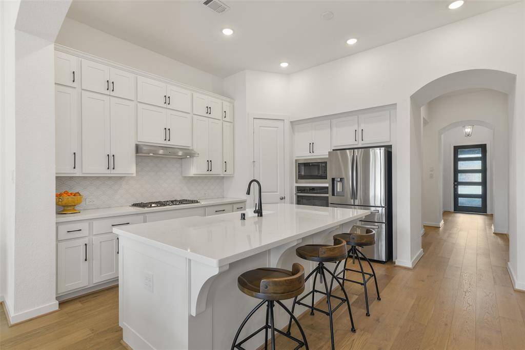 The island kitchen is a chef's dream, boasting all-white 42-inch cabinetry with stylish modern hardware, gleaming quartz countertops, added interest from an arabesque tile backsplash, engineered wood floors, and a high ceiling lit by recessed LED lighting.