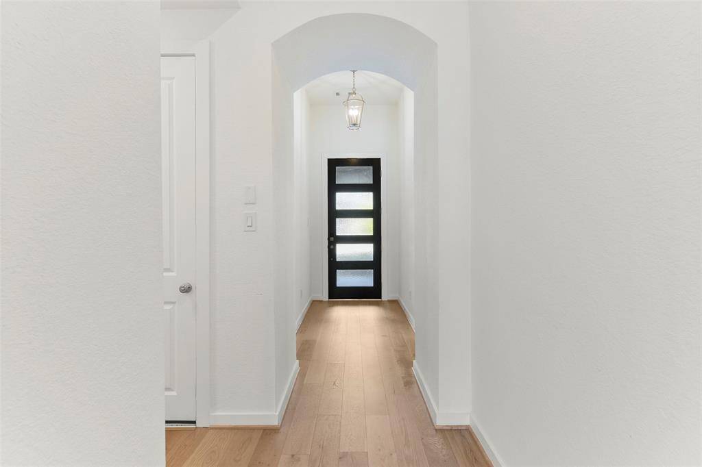 The modern-style front door with frosted glass panels opens to reveal a lengthy hallway with elevated ceilings, graceful arched doorways, neutral paint, engineered wood floors, and views all the way to the back of the home and beyond.