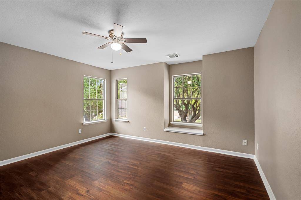 Your primary bedroom has laminate flooring and neutral walls, complemented by a ceiling fan. Three windows provide ample natural light and a view of the greenery outside.
