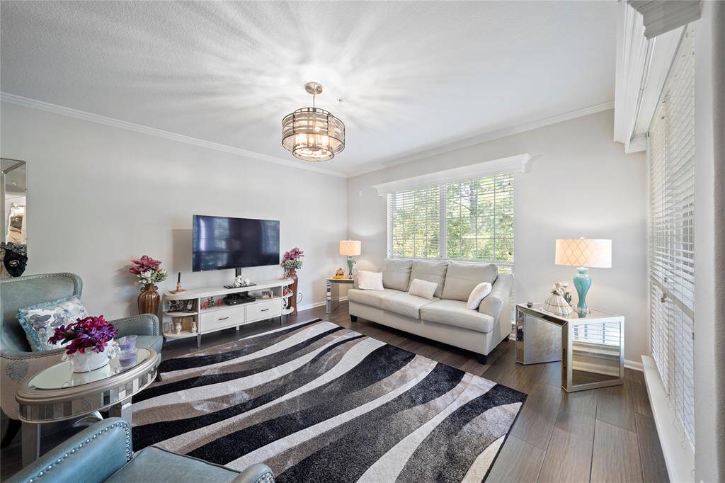 This living room features contemporary styling with a neutral color palette. Natural light filters through large windows with blinds, enhancing the room's airy feel. The space is well-appointed with comfortable seating, a modern ceiling light fixture, and a sleek TV setup, creating an inviting environment for relaxation and entertainment.