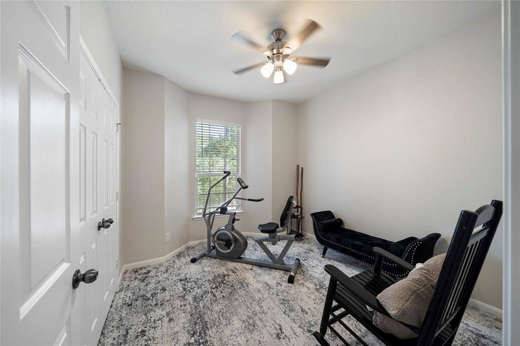 Versatile room featuring a ceiling fan, a large window for natural light, and neutral walls. It currently serves as a home gym with an exercise bike and weights, and has a cozy sitting area with a stylish black chair. The space can be easily personalized to fit your needs.
