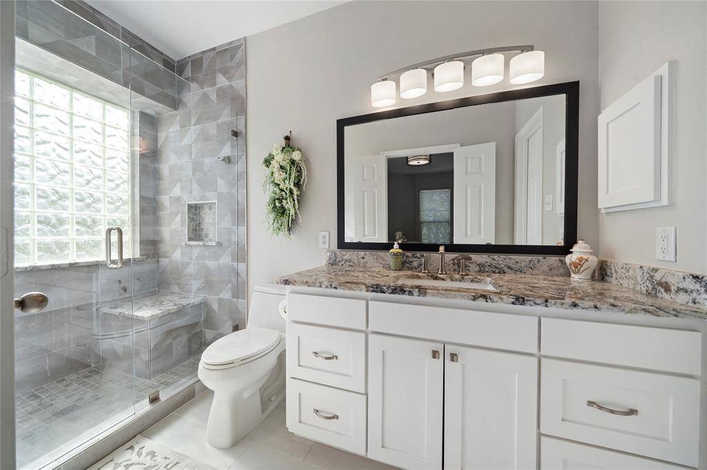 This is a modern bathroom featuring a spacious walk-in shower with glass doors, elegant gray tiling, a large window for natural light, a double vanity with granite countertops, and ample white cabinetry.