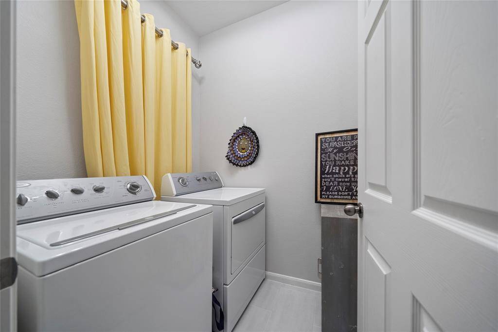 This is a compact and tidy laundry room with modern white appliances, including a washer and dryer, bright yellow curtains, and a cheerful decorative sign on the wall.