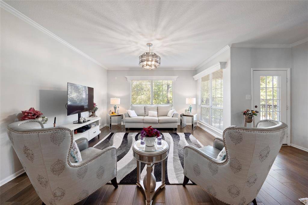 This is a well-lit, spacious living room featuring hardwood floors, elegant light fixtures, and large windows with blinds, leading to a cozy balcony.