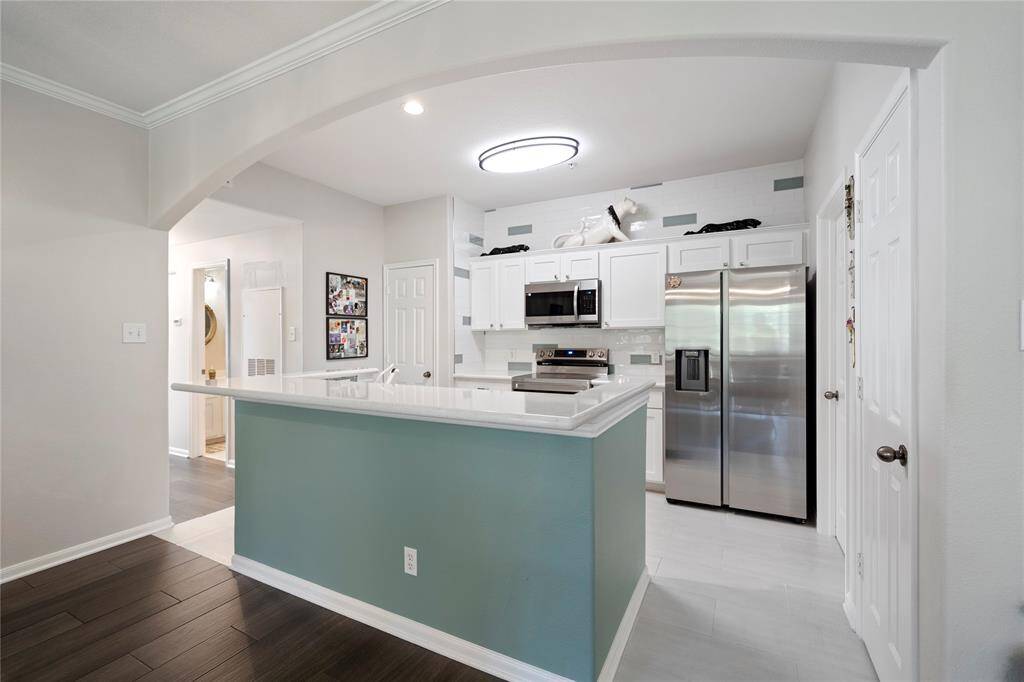 This is a bright, modern kitchen with white cabinetry, stainless steel appliances, and a contrasting island with a blue-green accent. The space is open and features a stylish arched opening, with wood flooring extending into the adjacent room.