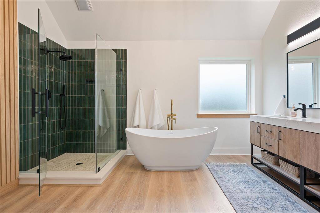 The spa-like walk-in shower features a striking dark green subway tile surround, laid in a sleek vertical pattern for a modern touch. A frameless glass enclosure enhances the open, airy feel. Just to the right, a freestanding soaking tub with sophisticated gold hardware invites you to relax in style.
