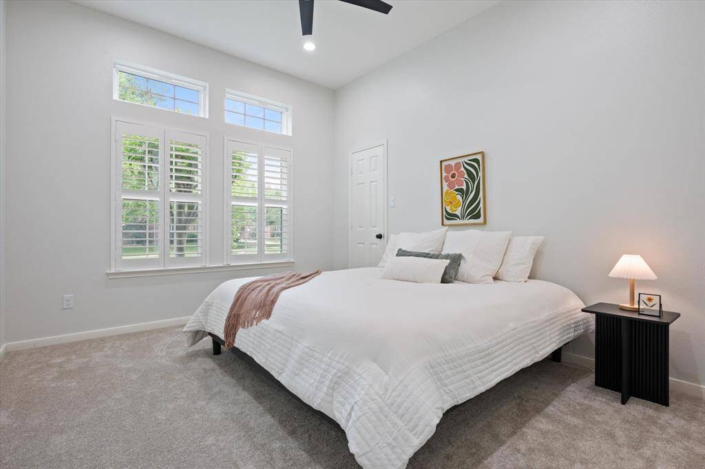 Guest bedroom number three is generously sized and features sophisticated decorator paint, a ceiling fan, and plush carpeting installed in 2025. Large windows, outfitted with plantation shutters, offer serene views of the front yard, enhancing the room's charm and natural light.