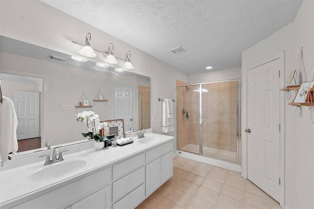 Primary bathroom features a double vanity with ample storage, a large mirror, and overhead lighting. It includes a spacious walk-in shower with glass doors and tile flooring. Neutral colors create a clean, bright atmosphere. Nice size linen closet and walk-in closet are also located within the primary bathroom as well. Lots of storage space!
