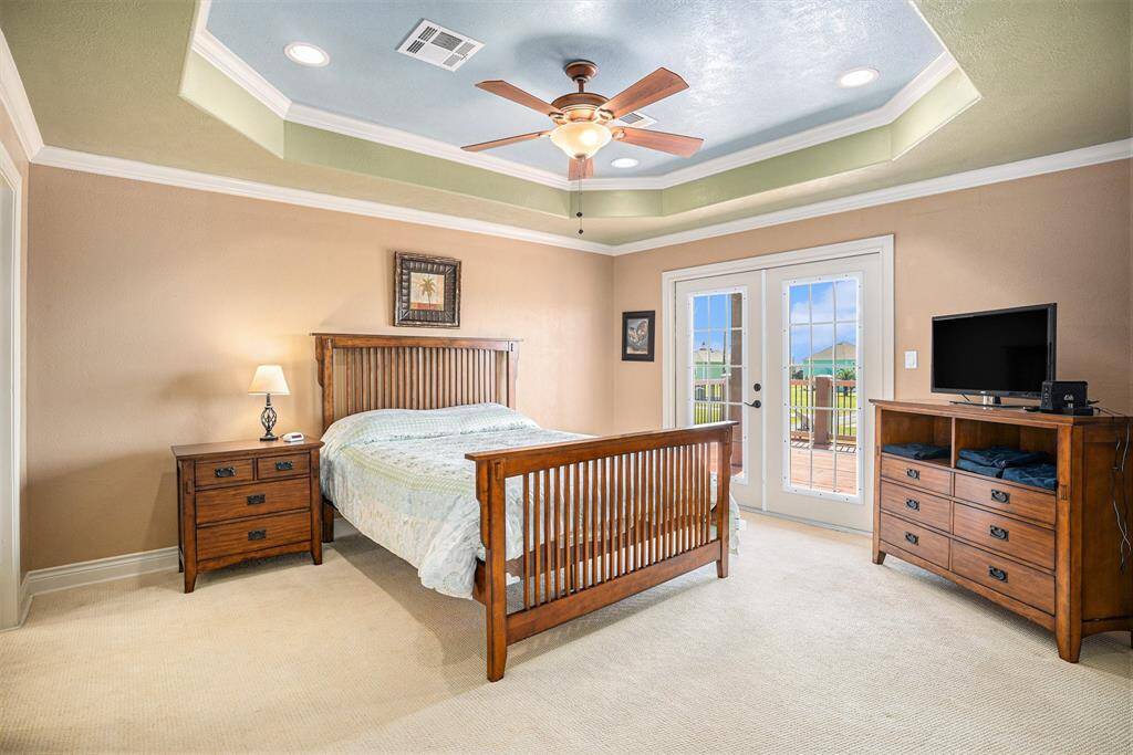 This spacious primary bedroom boasts a sophisticated tray ceiling and two sets of elegant French doors that open to the outside deck. The room is well-illuminated by a ceiling fan and recessed lighting, and is further enhanced by the refined touch of crown molding.
