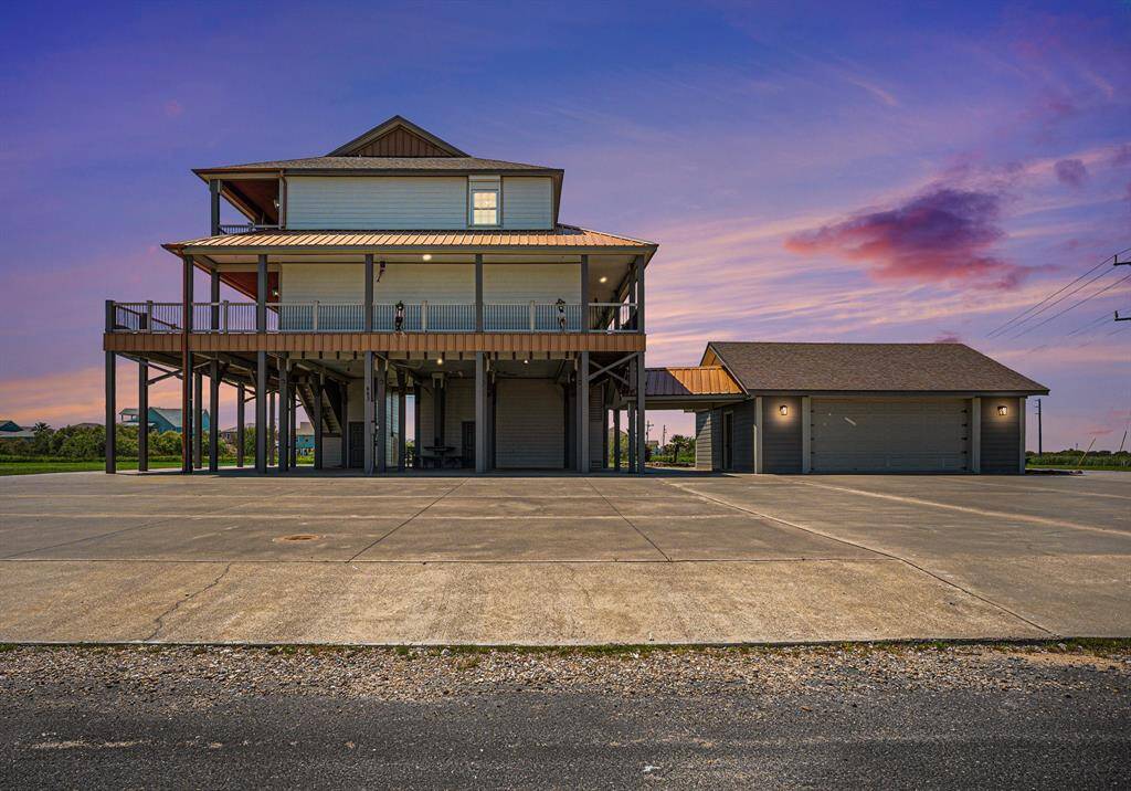 Stunning twilight view of elevated beach home with wraparound deck, spacious parking, and detached garage. Perfect for sunset relaxation.