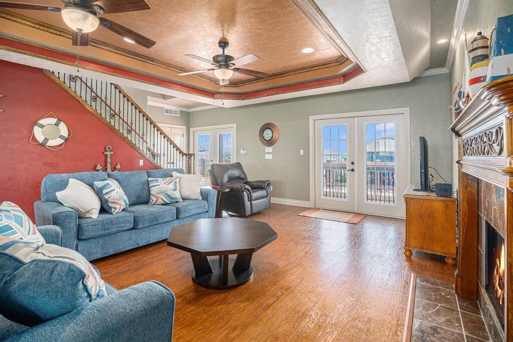 This bright and airy living room features a blend of coastal charm and comfortable living. The space is anchored by warm hardwood flooring and boasts a striking stone fireplace, adding a touch of rustic elegance. A vibrant red accent wall, adorned with nautical decor such as a life preserver and anchor, contributes to the room's character.  Double doors open to reveal a deck with expansive ocean views, seamlessly connecting indoor and outdoor living spaces and filling the room with natural light.