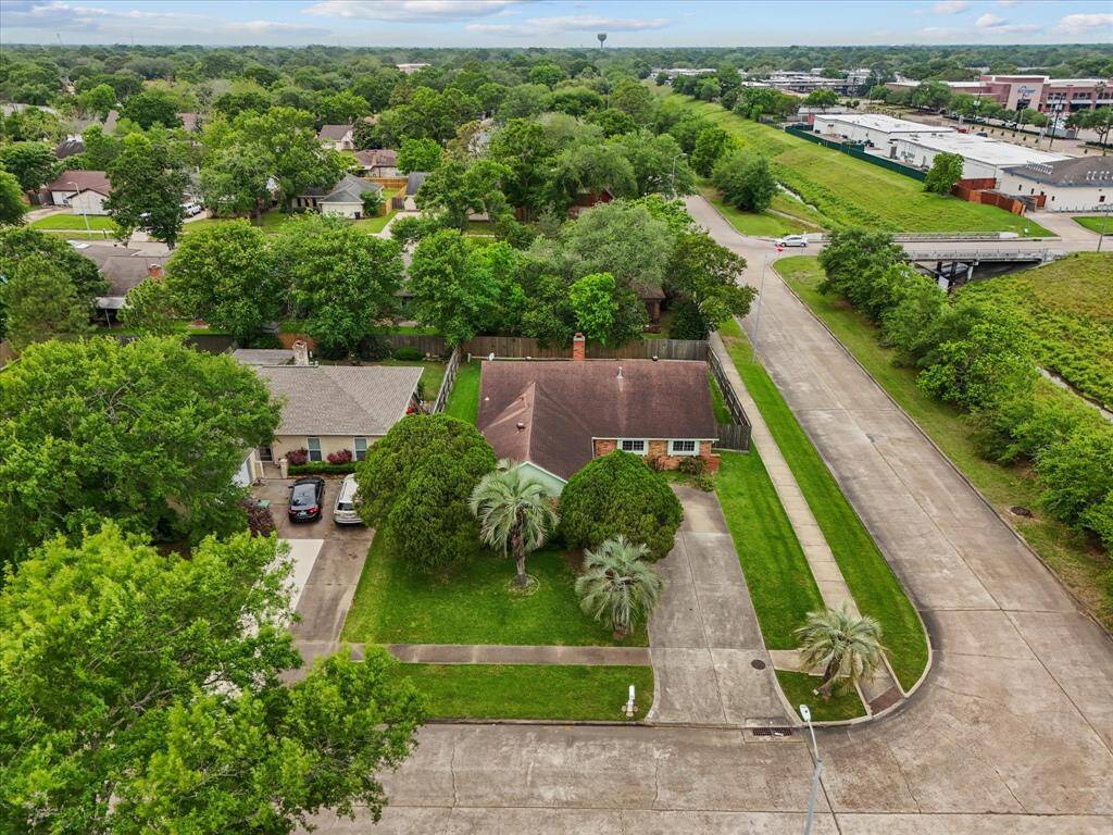 This aerial photo showcases a charming single-story home with a spacious yard, surrounded by lush greenery. It features a wide driveway, mature trees, and is situated in a quiet neighborhood with easy access to nearby amenities.