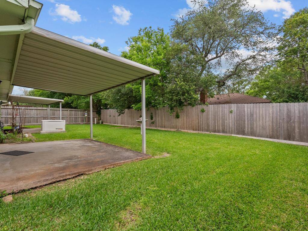 The photo shows a spacious backyard with a well-maintained lawn and a covered patio area. It features a wooden fence for privacy and is surrounded by mature trees, offering a serene outdoor space.