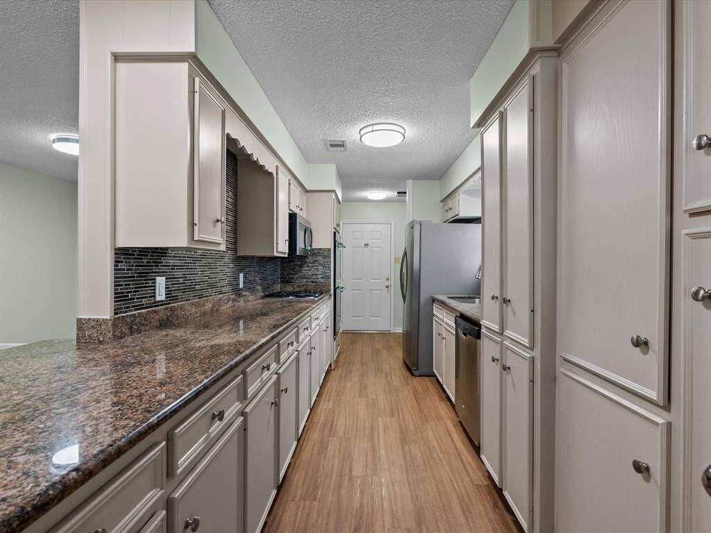 This kitchen features a galley layout with ample cabinetry, granite countertops, and a modern backsplash. It includes stainless steel appliances and wood flooring, offering a sleek and functional cooking space.