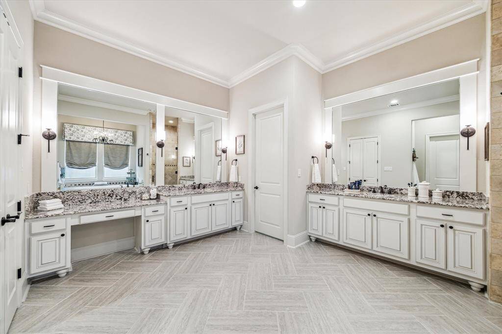 The luxurious ensuite features two separate vanities, each with granite countertops, framed mirrors, and elegant wall sconces that create a soft, flattering glow. Thoughtfully designed cabinetry offers abundant storage and counter space, along with convenient knee space to sit while you primp, blending beauty and function in perfect harmony.