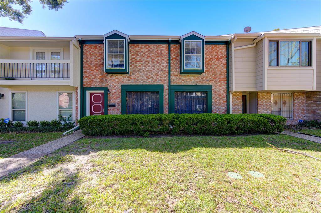 This charming two-story townhouse features a distinctive brick facade with green trim and two bay windows. A neatly landscaped front yard with shrubery enhances its curb appeal.