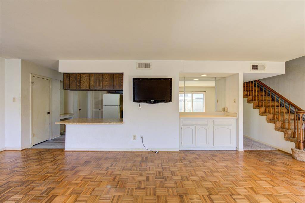 A view of the living area highlighting the parquet flooring and a breakfast bar opening into the kitchen. There is a built-in cabinet with a mirrored backsplash for added light and a carpeted staircase leading to the second floor.
