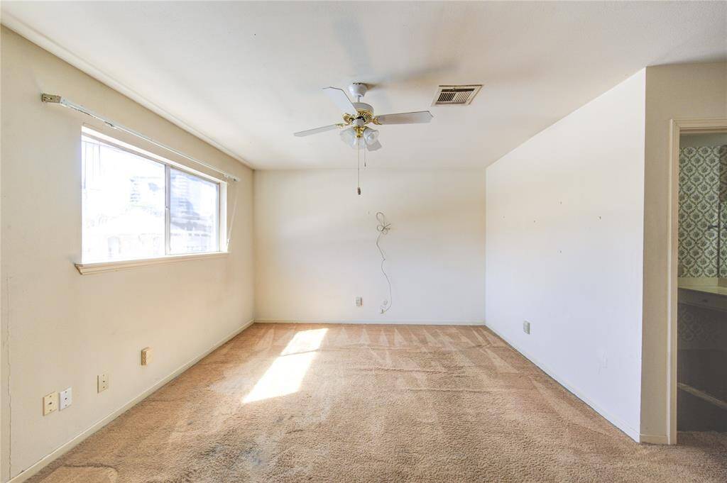 The bright primary bedroom with a large window, neutral walls, and carpet flooring. Features a ceiling fan and ample natural light.