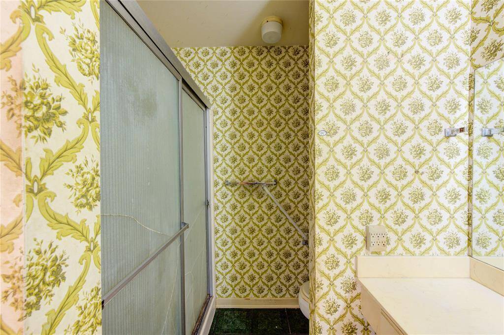 A vintage style bathroom with bold patterned wallpaper in green and white. It features a sliding glass shower door and a sink area. The space has a retro feel and will need updates.