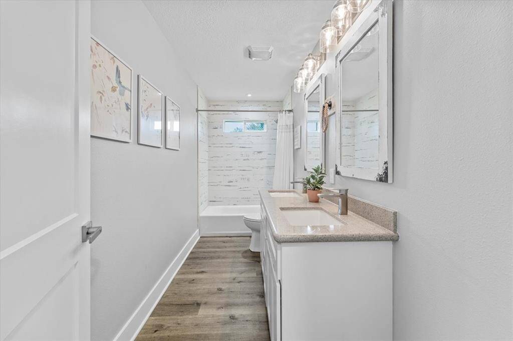This bathroom features a double vanity with modern fixtures, quartz countertops, two large mirrors, and bright lighting. The bright bathroom has wood-look flooring, and a shower-tub combo with window. Between each vanity is storage for all your personal items.