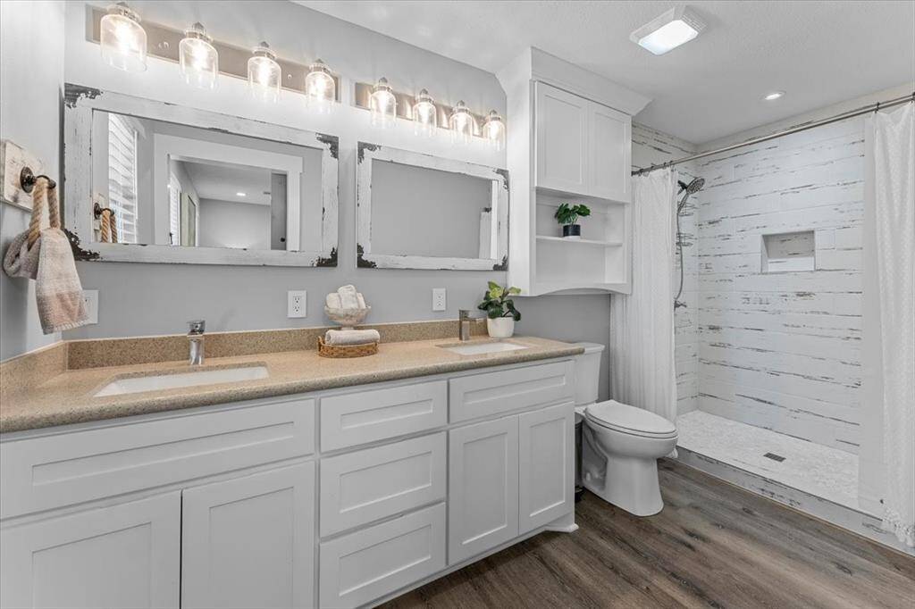 This bathroom features a modern design with dual sinks, a spacious vanity with ample storage, and stylish lighting fixtures. It includes a large walk-in shower with a rustic, tile wall, complemented by light quartz and wood look flooring for a warm, inviting feel.