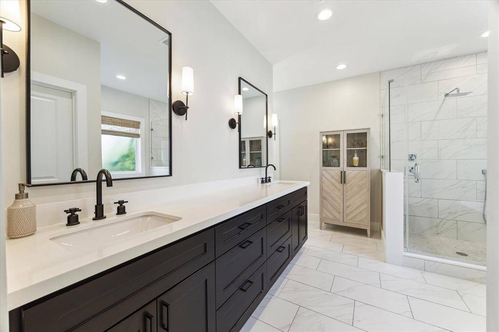 The ensuite bathroom offers a clean, modern aesthetic with contrasting tones and elevated finishes. Dual sinks sit beneath oversized mirrors, complemented by matching faucets and sconces. A spacious quartz-topped vanity provides ample storage, while the frameless glass shower features oversized marble-look tile and a sleek rainfall showerhead.