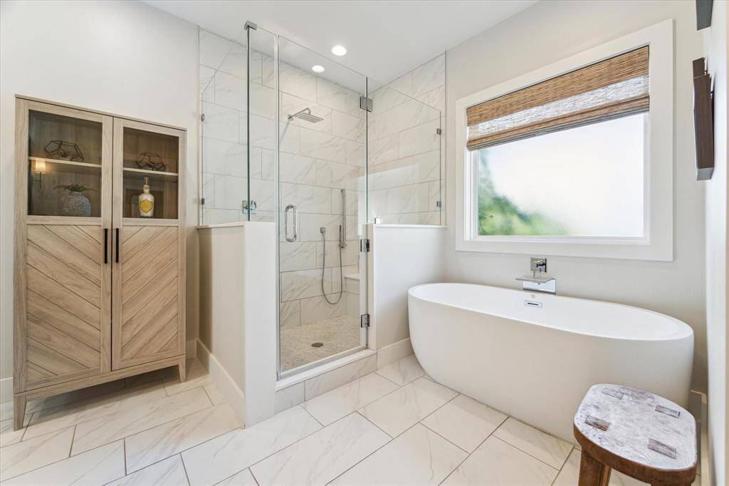 A freestanding soaking tub sits beneath a large frosted window, offering a peaceful spot to unwind. Nearby, the frameless glass shower showcases oversized marble-style tile, a built-in bench, and dual showerheads for a spa-like experience. Clean lines and natural light complete the serene and modern aesthetic.