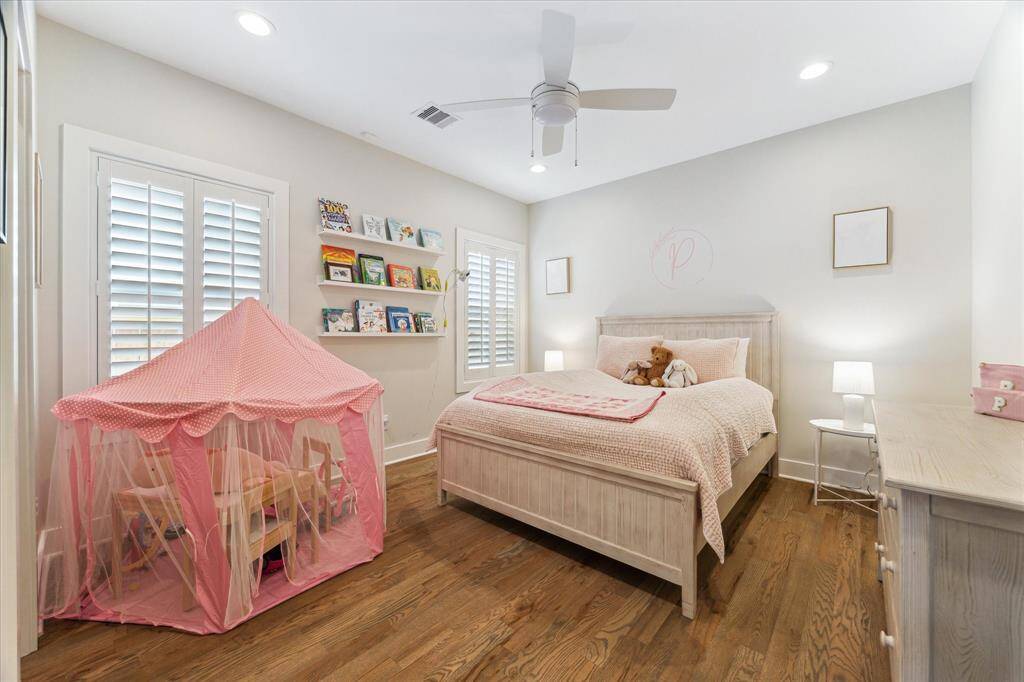This secondary bedroom features wide-plank hardwood floors, soft neutral walls, and plantation shutters that offer both privacy and natural light. Built-in shelving adds charm and storage, making it a playful yet peaceful space for rest or imagination.