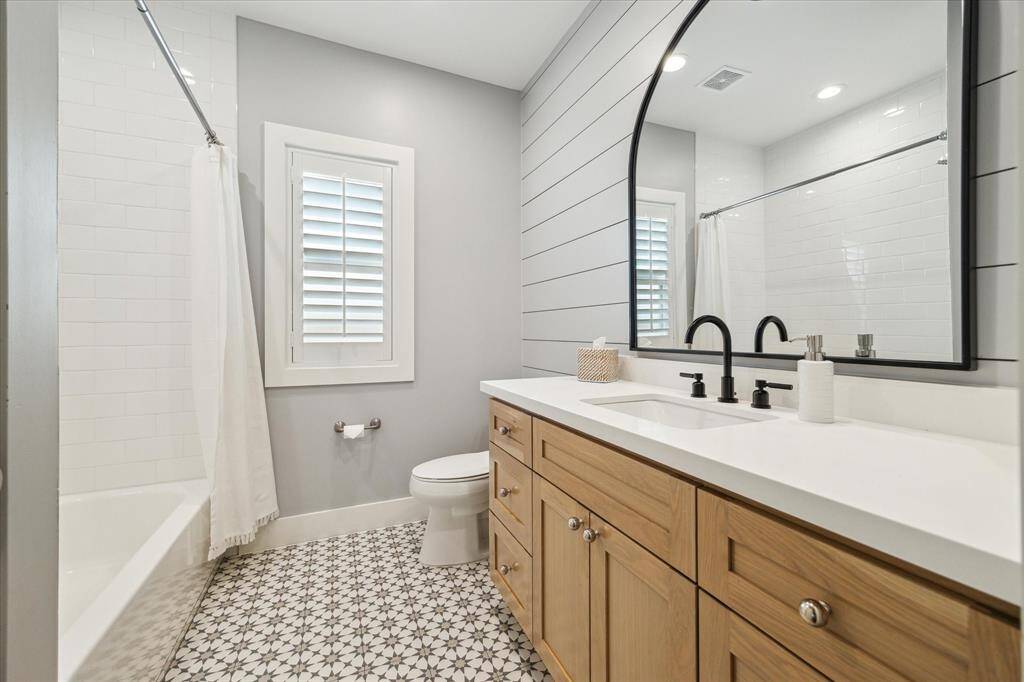 Patterned tile flooring, a wood-toned vanity, and a large arched mirror bring modern farmhouse charm to this secondary bathroom. Matte black fixtures contrast against the bright quartz countertop and white subway tile surround, while shiplap detailing and soft gray walls add texture and warmth.