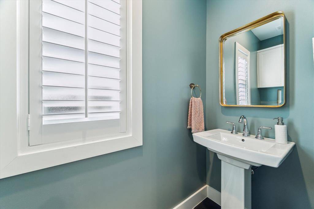 Located just off the mudroom, this half bath pairs a pedestal sink with polished chrome fixtures and a sleek, gold-framed mirror. Bold, moody walls contrast beautifully with crisp white trim and plantation shutters, giving the space a refined yet stylish feel.