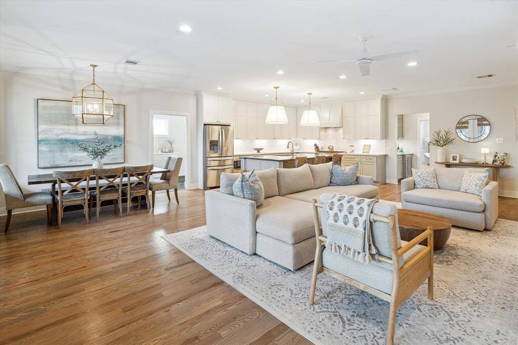 The open layout connects the living, dining, and kitchen areas with ease, creating a bright and functional gathering space. A gold statement chandelier anchors the dining area, while oversized pendants and sleek cabinetry give the kitchen a clean, modern edge.