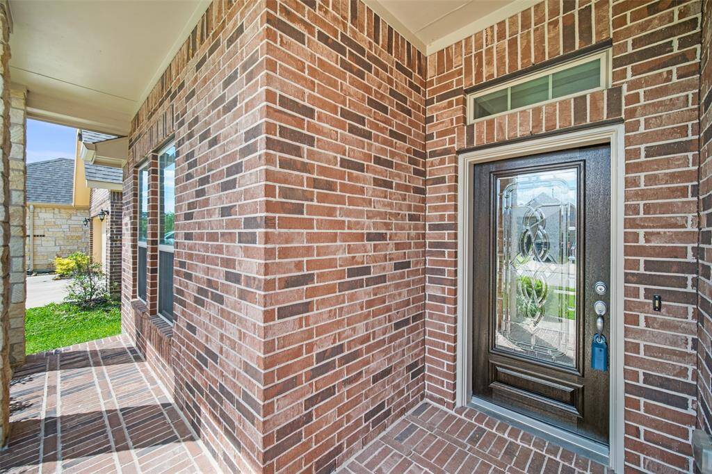 The covered front patio is framed with brick and stone column accents, complemented by a wooden front door with decorative glass etching, creating a warm and inviting entryway.