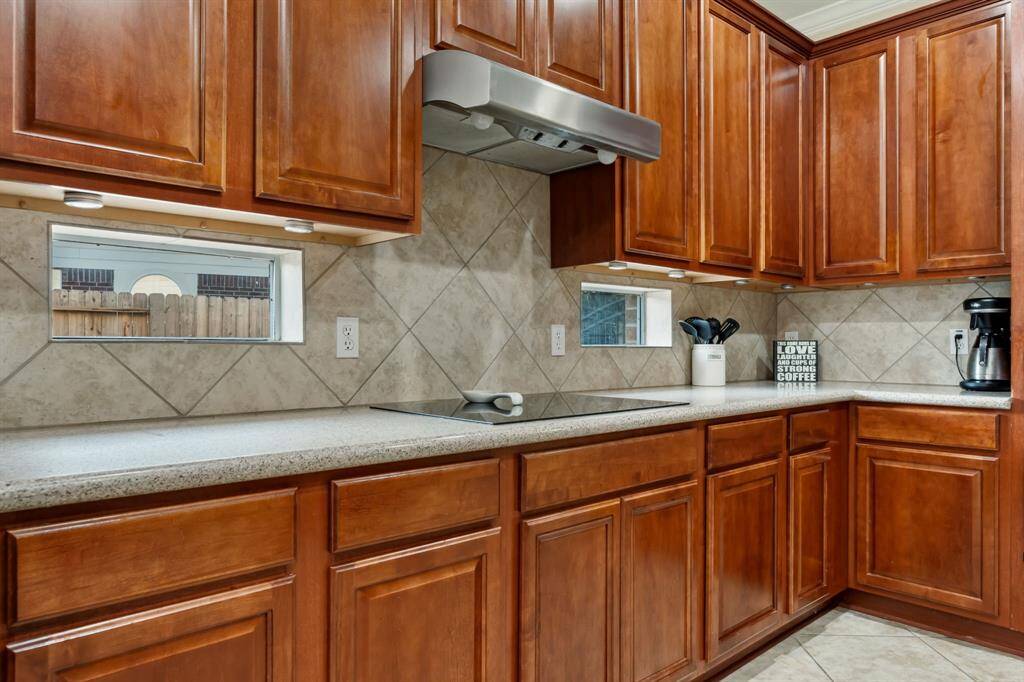 This kitchen features rich wooden cabinetry, a sleek cooktop, and a neutral-toned tile backsplash. Under-cabinet lighting, modern appliances, and small counter-level windows provide natural light.