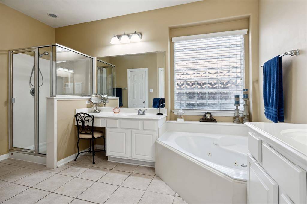 This bathroom features a spacious design with a glass-enclosed shower, a large jetted tub, and 2 vanities. Natural light fills the room through a sizable window, complementing the neutral tile flooring and walls.