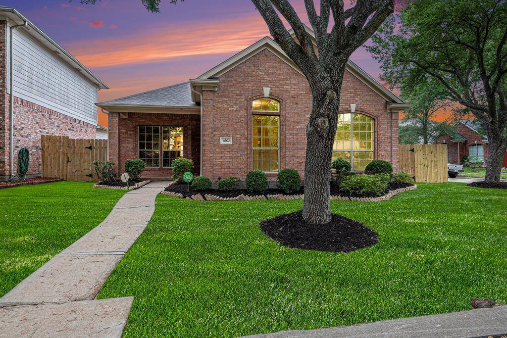 This charming brick home features a well-maintained front yard with lush green grass, mature trees, and neatly trimmed shrubs. A paved walkway leads to the inviting front porch, and large windows add to the home's curb appeal. The sunset backdrop enhances the cozy, welcoming atmosphere.