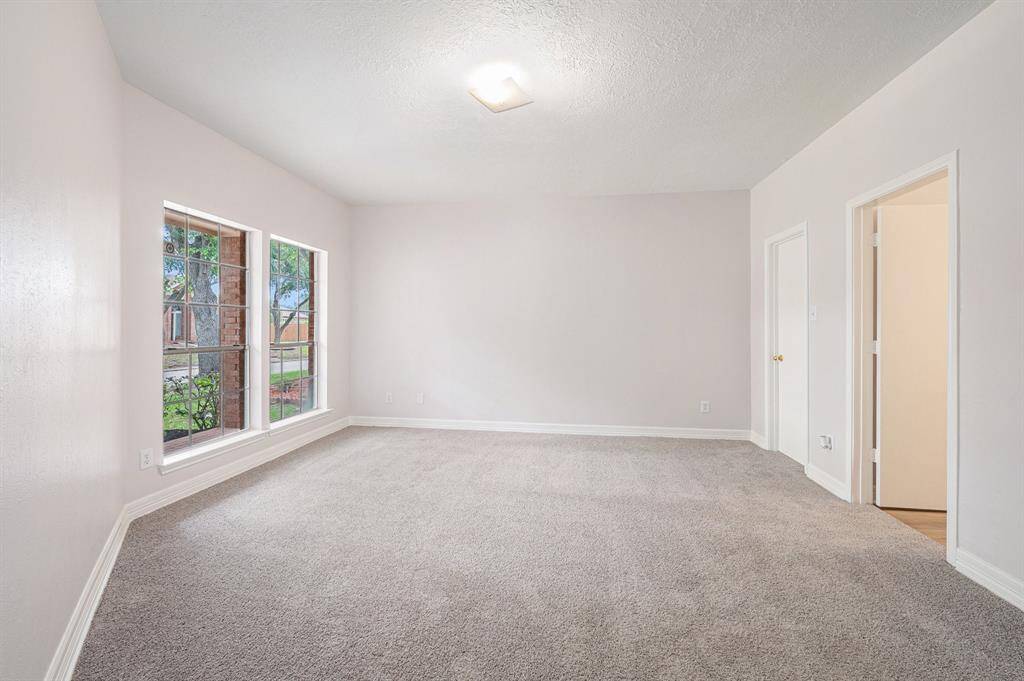 This primary room features neutral walls, plush carpeting, and large windows allowing natural light and neighborhood views. It offers a spacious, versatile layout with access to an adjoining area.