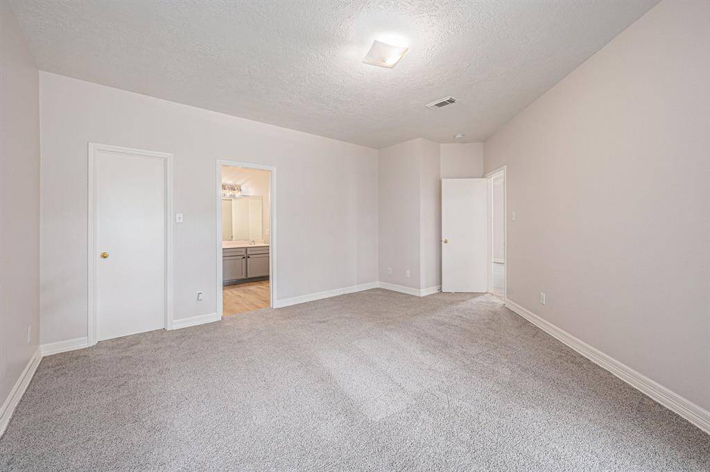 Spacious primary bedroom with neutral walls and carpet, featuring a ceiling light. It has two doorways, one leading to a bathroom with dual sinks and cabinetry, offering ample space and functionality.