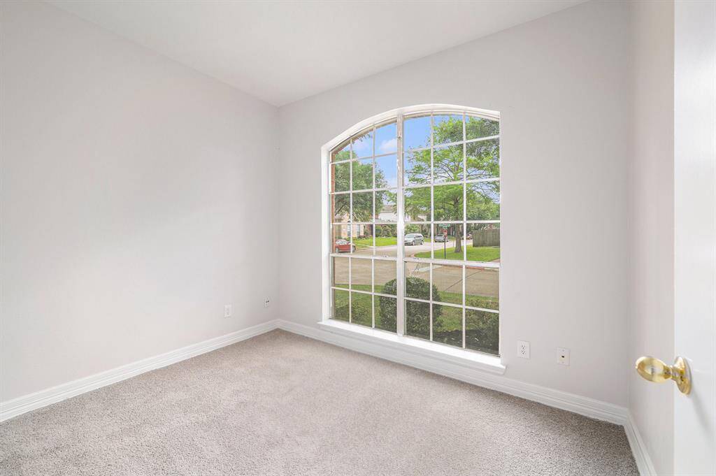 Bright and inviting room with neutral walls and plush carpet, featuring a large arched window offering plenty of natural light and a pleasant view of the street and greenery.