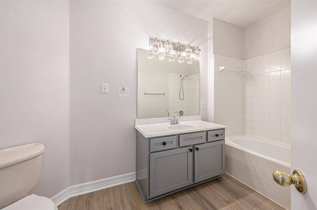 This bathroom features a modern design with a light gray vanity, a large mirror, and stylish lighting fixtures. It includes a bathtub with a classic white tile surround and wood-look flooring, creating a clean and inviting atmosphere.
