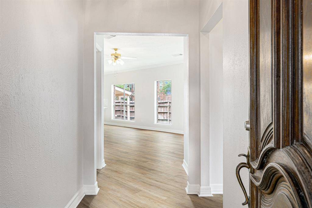 This photo shows a bright entryway with light wood flooring leading into a spacious, well-lit living area. The room has large windows that offer a view of the backyard, and a ceiling fan adds a touch of comfort.