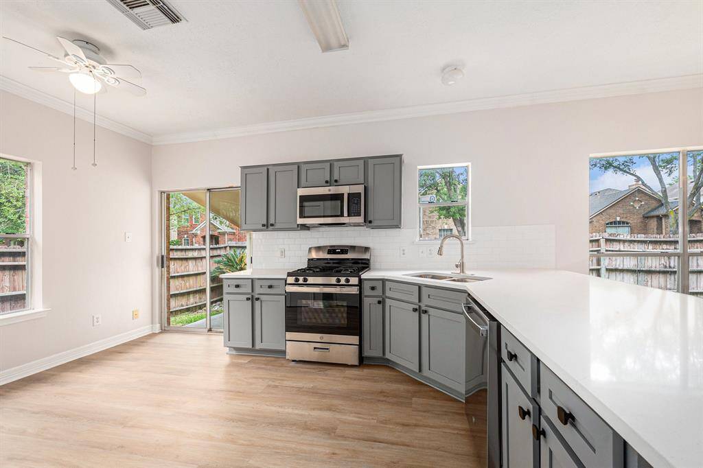 Bright and airy kitchen with light wood flooring, gray cabinets, and modern stainless steel appliances. Large windows and sliding glass door offer plenty of natural light.