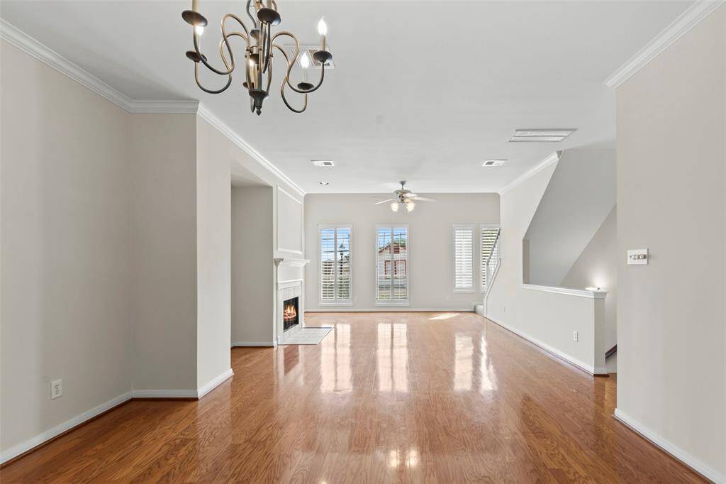 Bright and spacious living area with hardwood floors, a cozy fireplace, ceiling fan, and large windows with shutters. Elegant chandelier and neutral walls add a modern touch