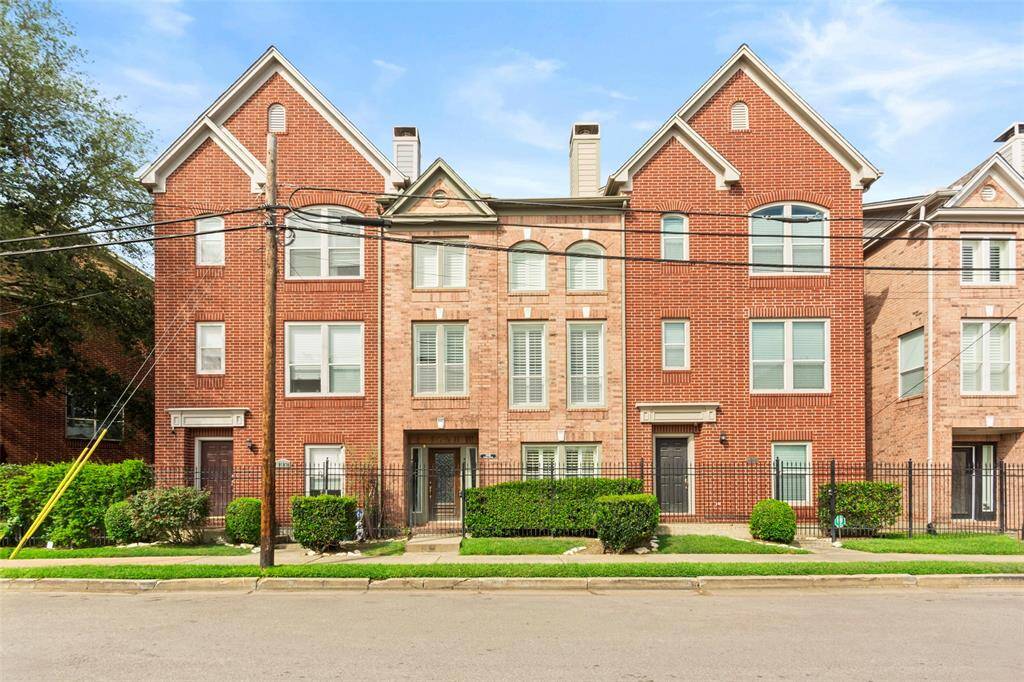 Modern red-brick townhouse with large windows and gable roofs. Features a fenced front yard, manicured shrubs, and a charming curb appeal