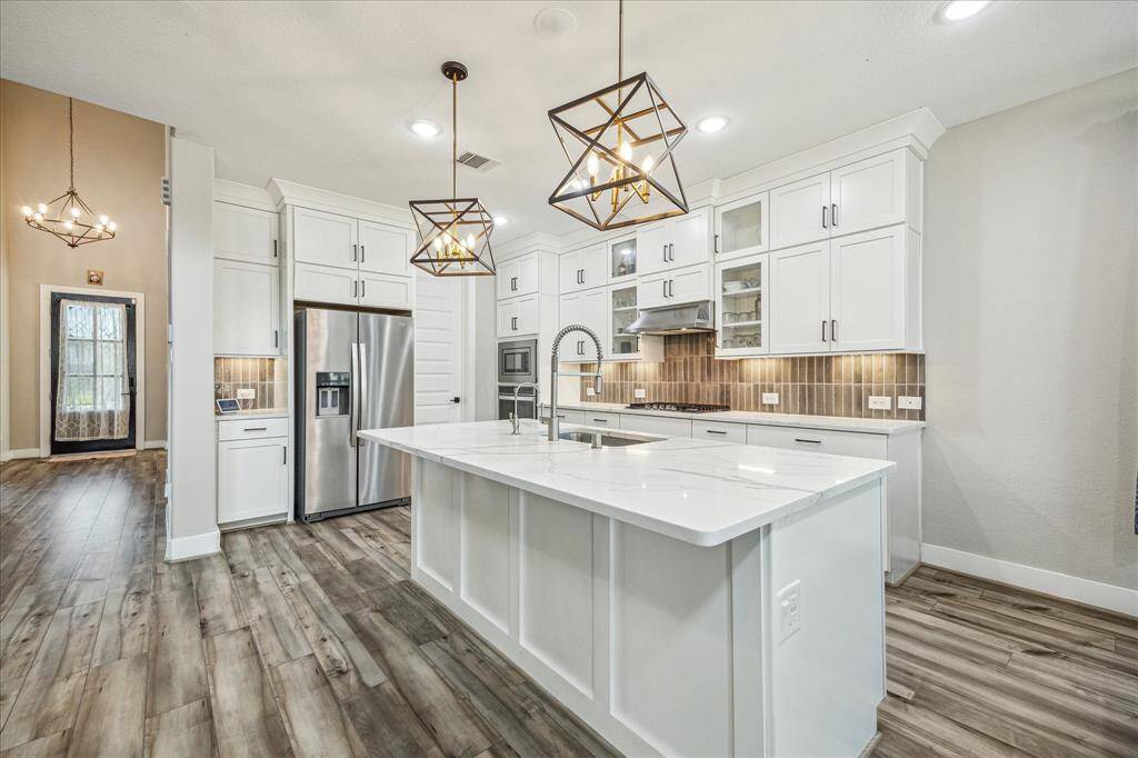 The kitchen showcases a spacious layout with a large island that includes an elegant waterfall-edge quartz countertop. Stainless steel appliances, a gas cooktop, and glass-front cabinetry add a sophisticated and functional touch to the modern design.