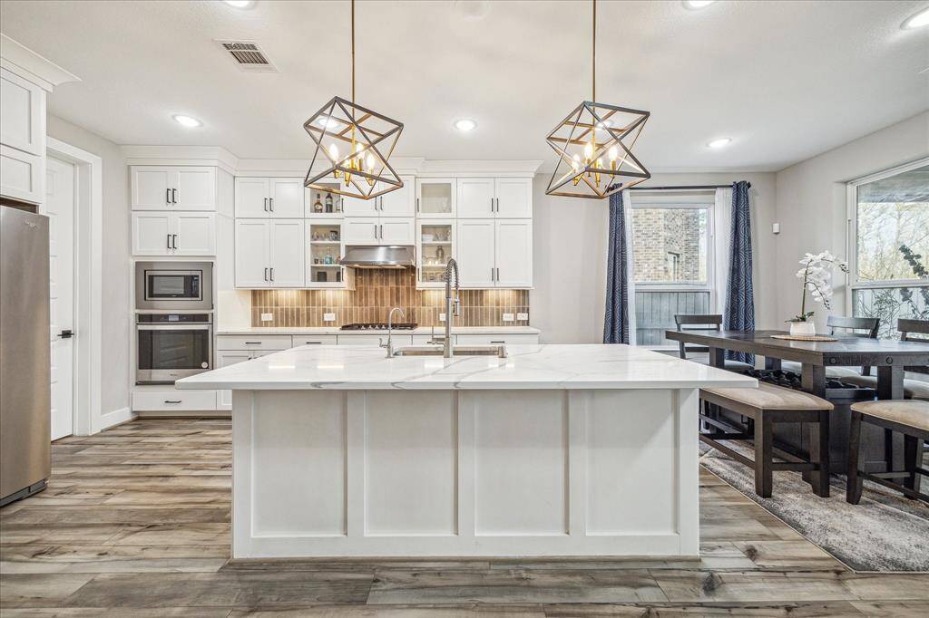The kitchen features a spacious island with a waterfall countertop and a sleek undermount sink, illuminated by two modern geometric pendant lights. The open layout seamlessly connects to the dining area, offering ample natural light through large windows and sliding glass doors.