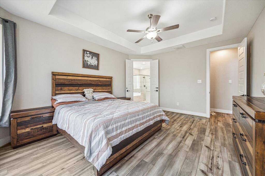 The primary bedroom features a tray ceiling with a ceiling fan, enhancing the spacious and airy feel of the room. Double doors lead to the en-suite bathroom, showcasing a seamless connection between the two spaces.
