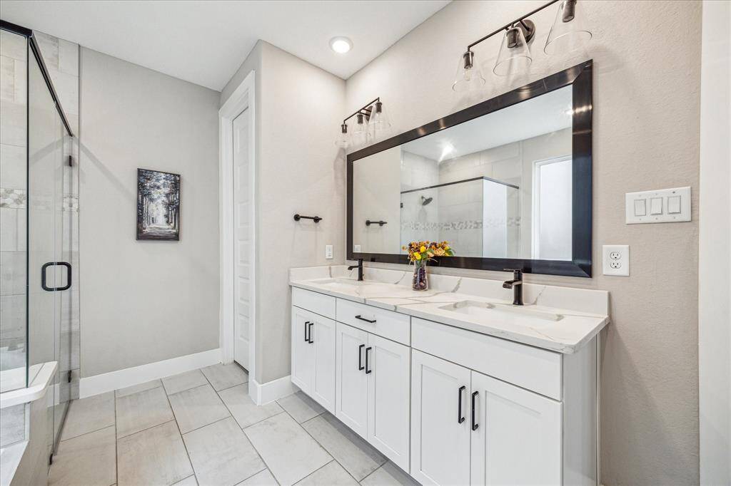 The primary bathroom features a modern double vanity with a sleek black-framed mirror and elegant light fixtures. A glass-enclosed shower is visible, complementing the neutral-toned tiles and contemporary finishes.